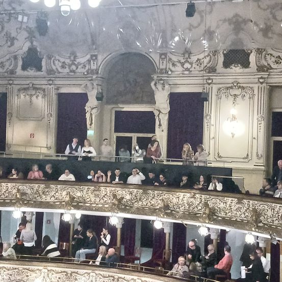 Ein volles Publikum in einem historischen Theater mit aufwendigen Balkonen und kunstvollen Verzierungen. Zuschauer sitzen auf roten Samtstühlen, einige stehen auf dem Balkon und schauen nach unten.