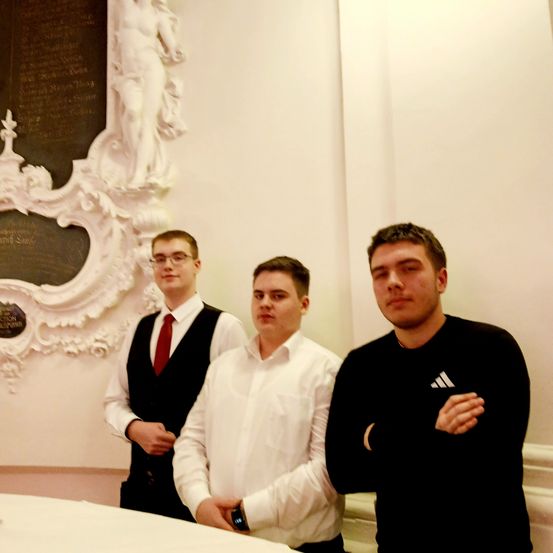 Drei Männer stehen vor einer dekorativen Wand, einer trägt eine rote Krawatte, ein anderer ein weißes Hemd und der dritte ein schwarzes Adidas-Shirt.