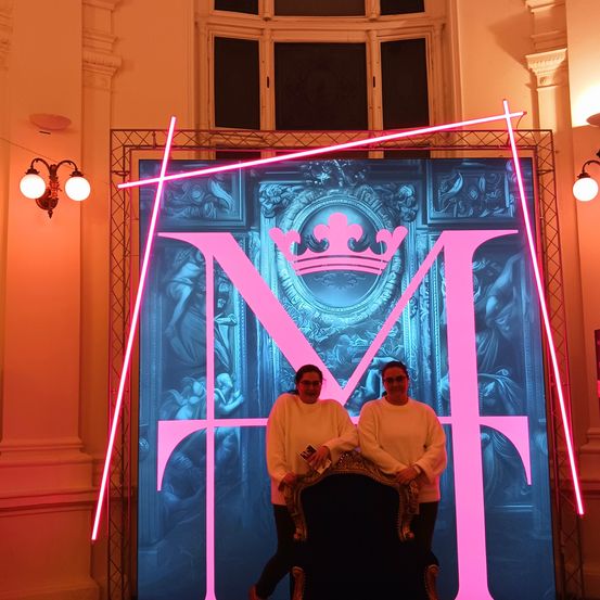 Zwei Personen stehen vor einem großen beleuchteten Bildschirm mit dem Buchstaben M und tragen weiße Langarmshirts. Dahinter kreuzt ein rosa Licht den Bildschirm, und eine große verzierte Tür ist abgebildet.