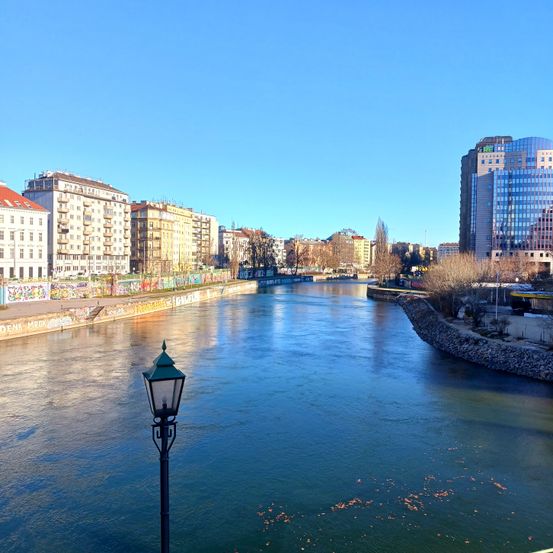 Ein Blick auf die Stadt mit einem Fluss, ruhigem Wasser, Gebäuden und Bäumen an einem klaren Tag. Ein Straßenlicht steht am Ufer des Flusses.