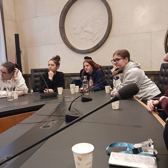 Fünf Personen sitzen um einen Tisch mit Mikrofonen herum, möglicherweise in einer Besprechung. Tassen, Flaschen und eine Tasche sind auf dem Tisch. Eine Person benutzt ein Handy. Der Raum hat ein kreisförmiges Wanddesign.