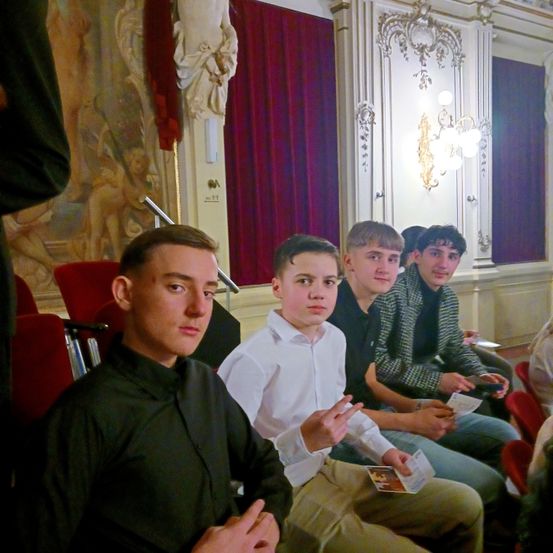 Fünf junge Männer sitzen in einer Reihe in einem Theater. Einer hält ein Stück Papier. Der Raum hat ein Gemälde und einen roten Vorhang.