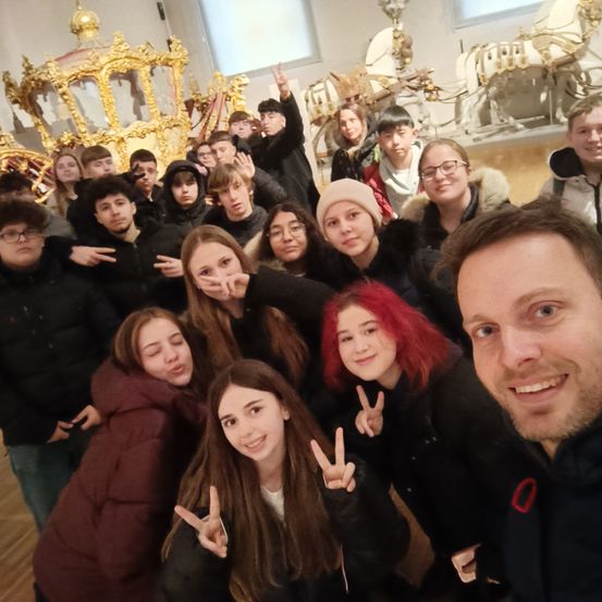Eine Gruppe junger Leute in Winterkleidung posiert für ein Foto in einem Museum mit einer goldenen Kutsche und Schlitten im Hintergrund.