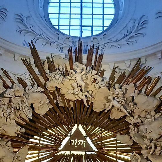 Ein goldenes kreisförmiges Emblem mit Strahlen umgibt eine dreieckige Form in der Mitte, geschmückt mit Cherubs und Wolken. Darüber befindet sich ein Fenster mit einem weißen Rahmen, das in eine dekorativ geschnitzte Decke eingelassen ist.