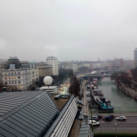 Luftaufnahme einer Stadt mit Gebäuden, Autos auf der Straße, einem Fluss und einer Brücke an einem nebligen Tag.