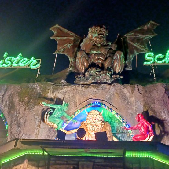 Eine Nachtszene zeigt eine große Statue eines Dämons auf einem felsigen Hügel mit Neonschildern. Unter der Statue befindet sich ein leuchtender Bogen, der mit bunten Lichtern geschmückt ist. Rechts ist ein roter Teufel und links eine grüne Kreatur zu sehen.