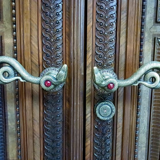 Ein Nahaufnahme von verzierten Holztüren mit Messinggriffen. Die Griffe haben ein detailliertes Schlangendesign und rote Edelsteine. Der Hintergrund ist kunstvoll geschnitztes Holz.