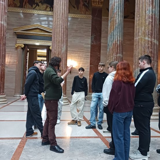 Ein Mann steht vor einer Gruppe von Menschen in einem großen, säulengestützten Saal und hält vermutlich einen Vortrag oder eine Präsentation.
