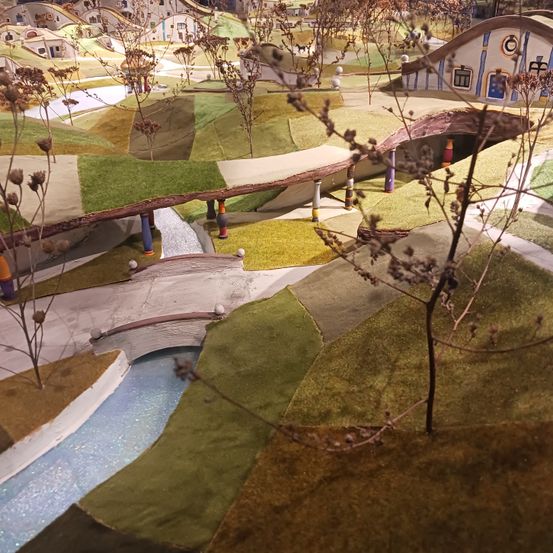 Eine Miniaturlandschaft mit kleinen Häusern, die in die Hänge gebaut sind und durch eine kleine Brücke über einen Fluss verbunden sind. Bäume und Sträucher sind verstreut.