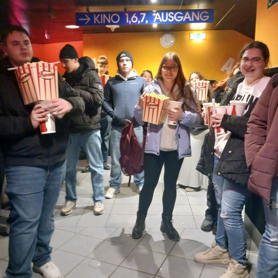 Eine Gruppe von Menschen steht in einem Flur mit Popcorn und Getränken, wahrscheinlich warten sie darauf, ein Kino zu betreten. Der Flur ist hell beleuchtet mit gelben Wänden und hat Schilder, die den Ausgang anzeigen.