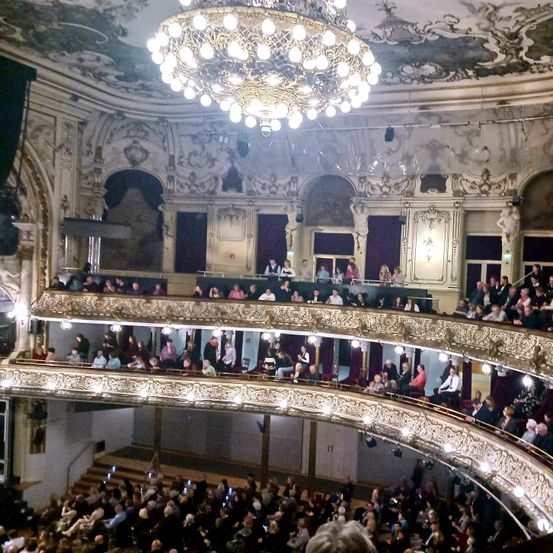 Ein Theater voller Menschen, die eine Aufführung beobachten. Die Bühne befindet sich in der Mitte, und darüber hängt eine große Kronleuchter. Das Publikum sitzt in Reihen auf den Balkonen.