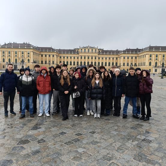 Eine Gruppe von Menschen steht vor einem großen Palast. Sie tragen Winterkleidung. Einige halten Taschen. Der Himmel ist bewölkt.