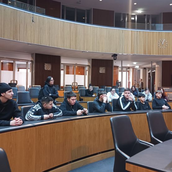 Eine Gruppe junger Menschen sitzt in einem Konferenzraum, einige mit den Händen im Gesicht, möglicherweise um einem Sprecher zuzuhören. Holztische, Stühle und eine Balkon mit einer Uhr sind im Hintergrund zu sehen.