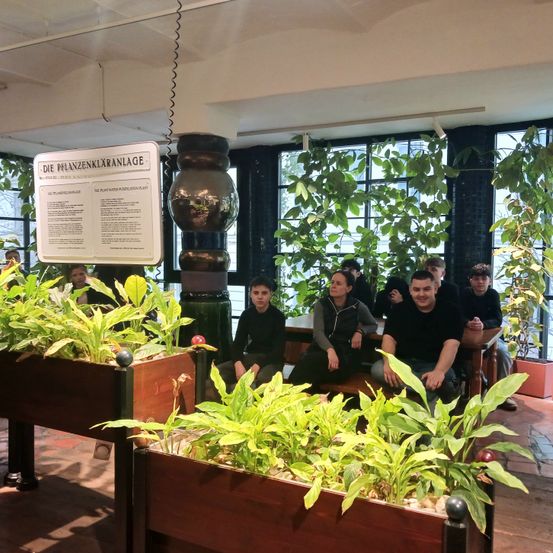 Eine Gruppe von Menschen sitzt in einem Gewächshaus, umgeben von Holzpflanzkübeln mit Grünpflanzen. Ein Schild mit der Aufschrift 'Die Pflanzenklaranlage' hängt darüber.