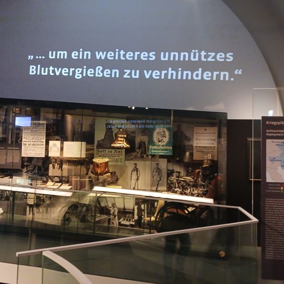 In einem Museumsaussteller sind verschiedene historische Gegenstände zu sehen, darunter Plakate und eine Trommel, unter einer Projektion, die '... um ein weiteres unnötiges Blutvergießen zu verhindern.' sagt.
