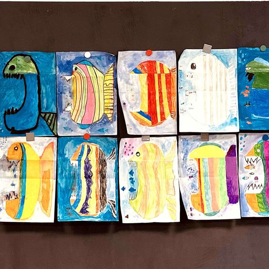 Eine Reihe bunter Fischgemälde ist an einer braunen Wand ausgestellt. Jedes Gemälde zeigt einzigartige Designs und Farben, und einige haben Papierclips angebracht.