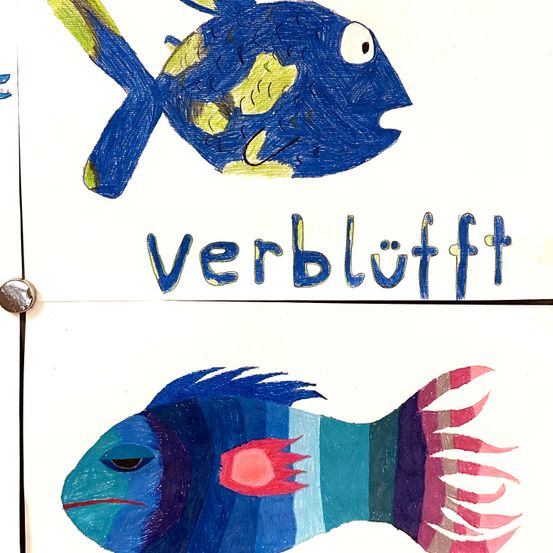 Zwei Zeichnungen von Fischen auf weißem Hintergrund. Der obere Fisch hat blaue Schuppen mit gelben Akzenten, ein weißes Auge und eine große Flosse. Darunter steht das Wort 'verblüfft' in Blau. Der untere Fisch ist bunt, mit blauen, grünen und roten Schuppen, einem roten Auge und einer langen Flosse.