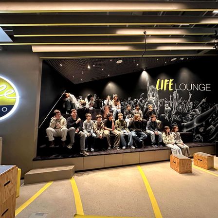 Eine Gruppe junger Menschen sitzt auf einer Bühne mit einem 'LIFE LOUNGE'-Wandbild dahinter. Sie sind von Holzkisten und einer gelben Linie auf dem Boden umgeben.