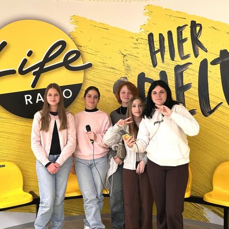 Fünf Frauen posieren vor einer gelben Wand mit 'Life Radio' Logo und 'Hier Beit' Text. Sie lächeln und halten ein Mikrofon.