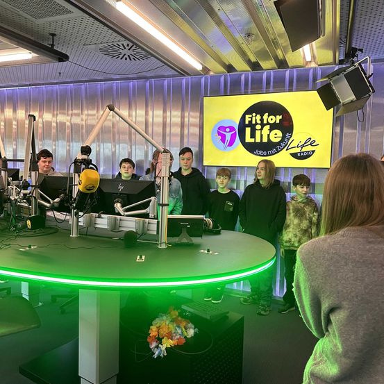 Eine Gruppe junger Menschen steht hinter einem Radio-Senderpult in einem Studio, mit einem gelben Banner, das 'Fit for Life' steht. Sie schauen in die Kamera, mit einer Frau in der Nähe.