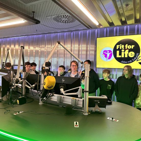 Eine Gruppe von Menschen steht hinter einem Tisch mit Mikrofonen, Monitoren und Lichtern. Der Raum hat ein gelbes Banner mit Fit for Life drauf.