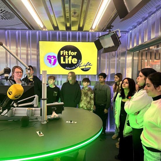 Eine Gruppe junger Menschen steht in einem Radiostudio und schaut auf ein Schild für Fit for Life. Eine Frau steht an einem Mikrofon, und andere stehen hinter ihr.