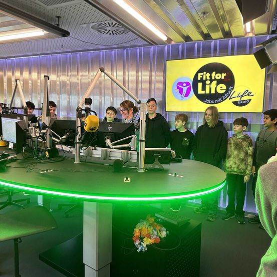 Eine Gruppe von Menschen steht hinter einem Schreibtisch in einem Radiostudio. Es gibt Computer, Mikrofone und ein Schild mit dem Text 'Fit for Life' an der Wand.