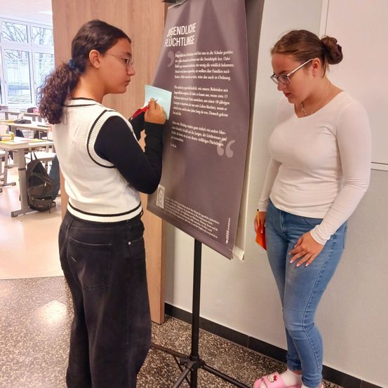 Zwei Frauen stehen in einem Klassenzimmer neben einem Plakat. Eine schreibt auf das Papier, während die andere sie ansieht.