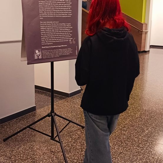 Eine Person mit roten Haaren schaut auf ein Display-Board mit Text vor einer weißen Wand.