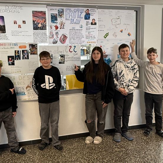 Fünf junge Personen stehen gemeinsam vor einer Pinnwand, lächeln und posieren für ein Foto. Die Tafel ist mit verschiedenen farbenfrohen Postern und Zeichnungen gefüllt.