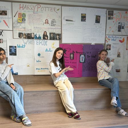 Drei Mädchen sitzen auf einer Holztreppe und lächeln für ein Foto. Hinter ihnen hängen Poster mit verschiedenen Themen, darunter Harry Potter, Whitefox und ein vampirthematisches Poster.