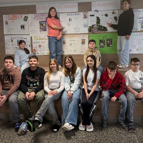 Eine Gruppe von Schülern posiert für ein Foto in einem Klassenzimmer mit Postern an der Wand. Ein Schüler steht in der Mitte.