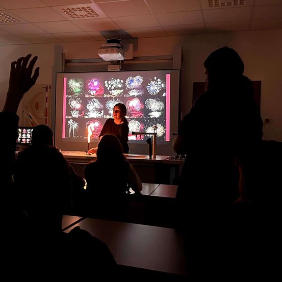 Eine Frau unterrichtet eine Klasse mit einem Projektor, der Feuerwerke anzeigt, während sie eine Kerze hält. Die Schüler sehen von ihren Plätzen zu.
