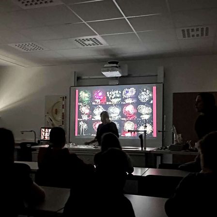 Ein Klassenzimmer mit einer Projektionsleinwand, die Bilder von Feuerwerken zeigt. Eine Person steht vor der Leinwand. Mehrere Schüler sitzen an Tischen.