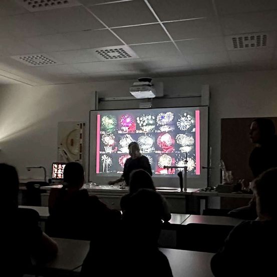 Ein Klassenzimmer mit einer Projektionsleinwand, die Bilder von Feuerwerken zeigt. Eine Person steht vor der Leinwand. Mehrere Schüler sitzen an Tischen.