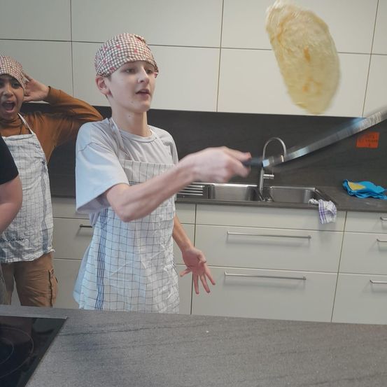 Ein junger Mensch in der Küche wirft eine Tortilla in die Luft, während eine andere Person dahinter überrascht zu sein scheint. Sie tragen Schürzen und stehen in der Nähe einer Spüle.