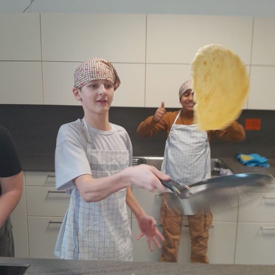 Zwei Personen befinden sich in einer Küche, eine flippt eine Tortilla, während eine andere Daumen hoch hält. Beide tragen Schürzen, und eine dritte Person steht in der Nähe. Die Küche hat weiße Schränke und eine Arbeitsplatte.