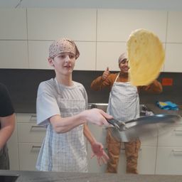 Zwei Personen befinden sich in einer Küche, eine flippt eine Tortilla, während eine andere Daumen hoch hält. Beide tragen Schürzen, und eine dritte Person steht in der Nähe. Die Küche hat weiße Schränke und eine Arbeitsplatte.