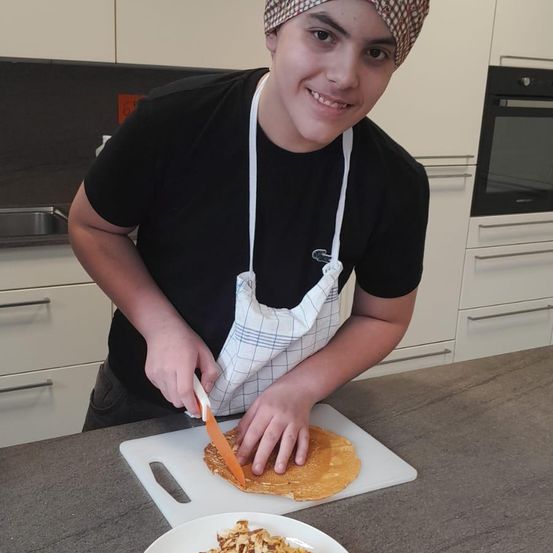 Ein junger Junge mit einem karierten Kopftuch und einer Schürze schneidet eine Tortilla auf einem Schneidebrett, neben einem Teller mit gehackten Nüssen. Er lächelt und scheint die Aktivität zu genießen.