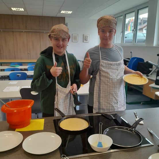 Zwei Jungen stehen in einer Klassenküche, tragen Schürzen und halten gekochte Crêpes auf einem Teller. Ein Junge gibt ein Daumen hoch. Der Tresen hat mehrere Teller, Pfannen und eine Schüssel.