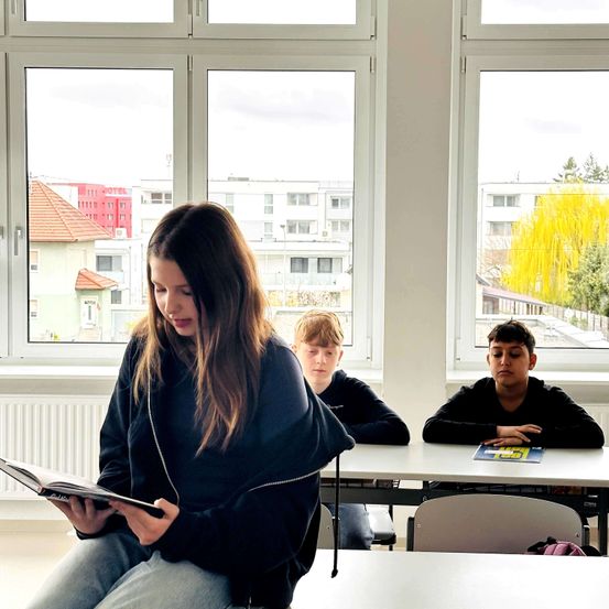 Drei Schüler sitzen in einem Klassenzimmer mit großen Fenstern, die eine Stadtlandschaft überblicken. Das Mädchen vorne liest ein Buch, während die beiden Jungen hinter ihr die Arme verschränkt haben.