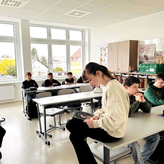 Ein Klassenzimmer mit Schülern an Tischen. Ein Mädchen liest ein Buch, während andere zuhören. Fenster und Bücherregale sind sichtbar.