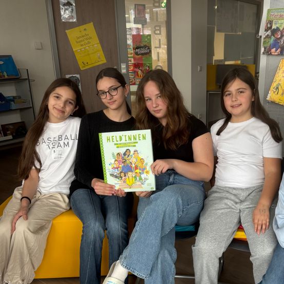 Vier Mädchen sitzen auf einer gelben Bank und lächeln. Die mittlere hält ein Buch mit dem Titel Heldinnen. Dahinter befindet sich ein Poster und ein Regal mit Büchern.