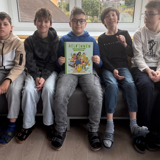 Fünf Jungen sitzen auf einer Bank, einer von ihnen hält ein Comic-Buch mit dem Titel 'Heldinnnen.' Sie tragen Freizeitkleidung und schauen zur Kamera. Hinter ihnen zeigt ein Fenster ein Haus mit Bäumen.