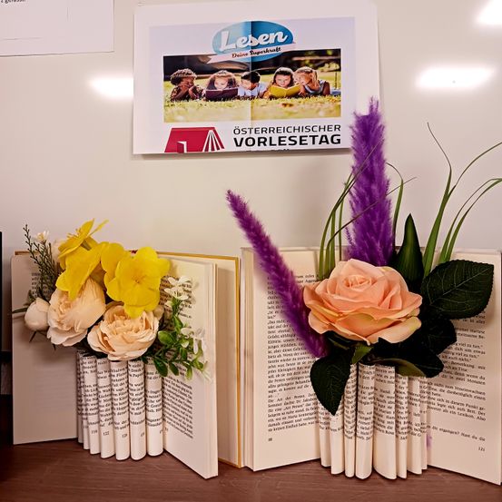 Zwei offene Bücher, dekoriert mit Blumen und Blättern auf einem Tisch. Hinter den Büchern ist ein Plakat mit dem Text 'Österreichischer Vorlesetag'.