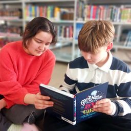 Ein junges Mädchen und ein Junge sitzen auf dem Boden in einer Bibliothek und lesen ein Buch mit dem Titel 'Gute Nacht Geschichten für Rebellische Mädchen'. Sie sind von Bücherregalen umgeben.