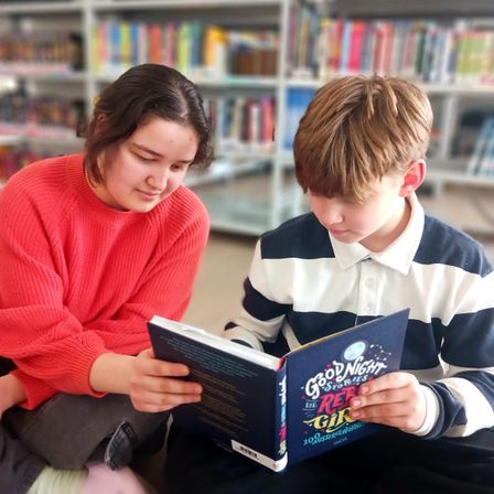 Ein junges Mädchen und ein Junge sitzen auf dem Boden in einer Bibliothek und lesen ein Buch mit dem Titel 'Gute Nacht Geschichten für Rebellische Mädchen'. Sie sind von Bücherregalen umgeben.