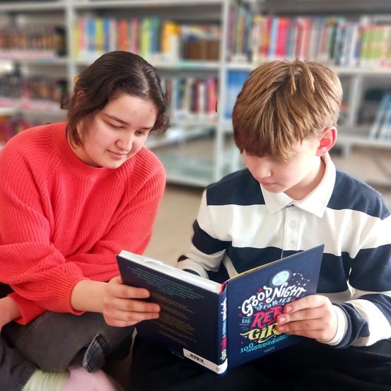 Ein junges Mädchen und ein Junge sitzen auf dem Boden in einer Bibliothek und lesen ein Buch mit dem Titel 'Gute Nacht Geschichten für Rebellische Mädchen'. Sie sind von Bücherregalen umgeben.