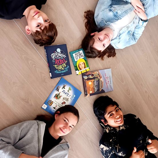 Vier Kinder liegen auf einem Holzboden, jedes hält ein Buch. Sie scheinen für ein Foto zu posieren. Ein Mädchen lächelt in die Kamera.