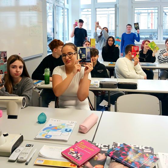 Ein Klassenzimmer mit Schülern in Reihen, die Bücher halten. Eine Frau in Weiß steht vorne und hält ein Buch. Auf dem Tisch liegen andere Bücher, eine Fernbedienung und ein Stift.